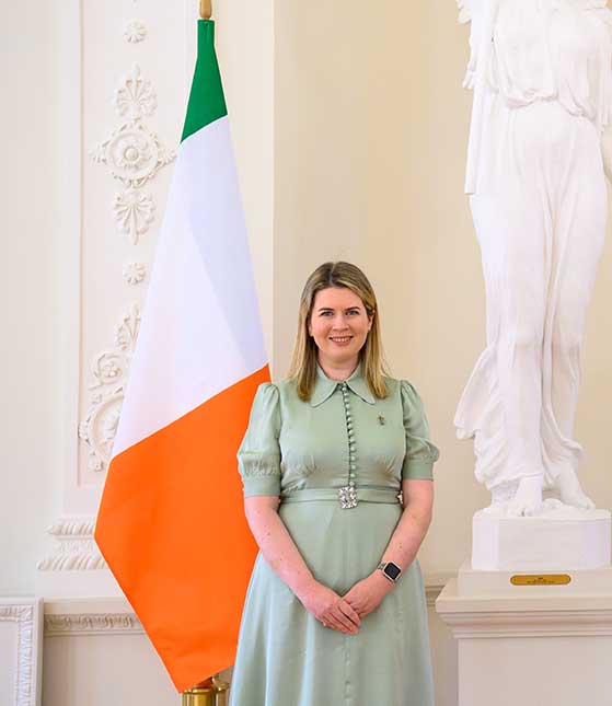 Ambassador Seadhna MacHugh wearing a green dress, standing in front of the Irish flag.